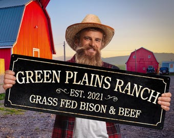 May include: A black metal sign with gold lettering that reads "GREEN PLAINS RANCH EST. 2021 GRASS FED BISON & BEEF". The sign is held in front of a red barn and a person wearing a straw hat.