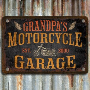 Pu&ograve; includere: Cartello in metallo rustico con il testo "Garage della Moto di Nonno" in lettere arancioni e nere. Il cartello presenta anche un'illustrazione di una moto d'epoca e il testo "Est. 2000".