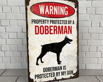 May include: A white metal sign with a red warning sign that says "WARNING" and the text "PROPERTY PROTECTED BY A DOBERMAN. DOBERMAN IS PROTECTED BY MY GUN". A black silhouette of a Doberman Pinscher is in the center of the sign.