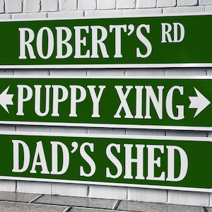 May include: Three green metal signs with white lettering. The top sign reads "Robert's Rd". The middle sign reads "Puppy Xing" with an arrow pointing left and an arrow pointing right. The bottom sign reads "Dad's Shed".