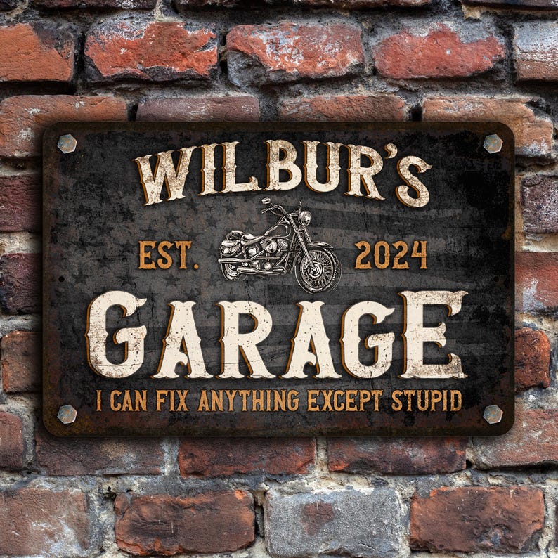 May include: Black metal sign with white text that reads "Wilbur's Garage Est. 2024 I can fix anything except stupid". The sign features a vintage motorcycle graphic.