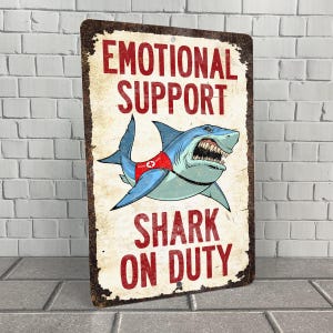 May include: A vintage-style metal sign with a cartoon shark wearing a red lifeguard vest. The sign reads "Emotional Support Shark On Duty".