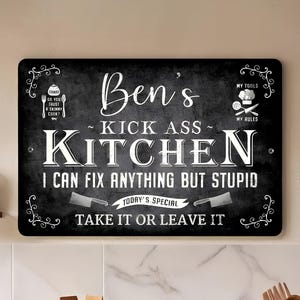 May include: Black rectangular metal sign with white text that reads "Ben's Kick Ass Kitchen". The sign includes the phrases "I can fix anything but stupid" and "Take it or leave it". Decorative flourishes are in each corner.