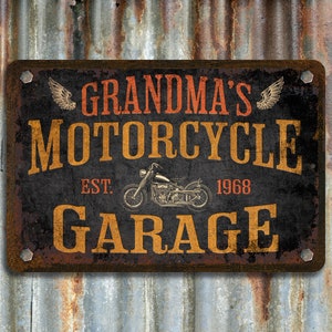 Pu&ograve; includere: Un cartello di metallo arrugginito con il testo "Grandma's Motorcycle Garage" in lettere arancioni e gialle. Il cartello include anche il testo "Est. 1968" e un'immagine in bianco e nero di una motocicletta.