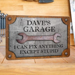 Puede incluir: Un letrero met&aacute;lico rectangular con un acabado envejecido. El letrero dice "DAVE'S GARAGE" encima de un gr&aacute;fico de llave oxidada y el texto "I CAN FIX ANYTHING EXCEPT STUPID."