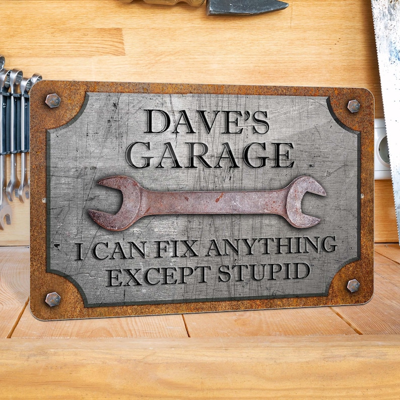 Puede incluir: Un letrero met&aacute;lico rectangular con un acabado envejecido. El letrero dice "DAVE'S GARAGE" encima de un gr&aacute;fico de llave oxidada y el texto "I CAN FIX ANYTHING EXCEPT STUPID."