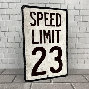 May include: A vintage-style metal sign with a white background and black text. The sign reads "SPEED LIMIT 23".