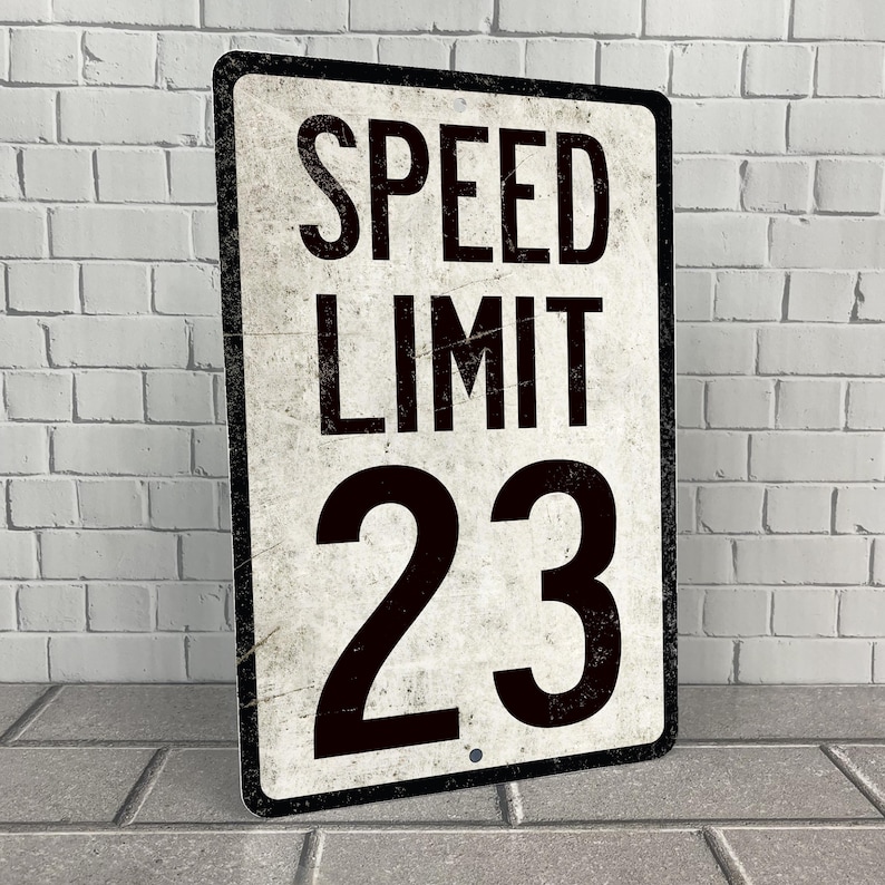 May include: A vintage-style metal sign with a white background and black text. The sign reads "SPEED LIMIT 23".