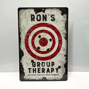 May include: A white metal sign with a red and white target with bullet holes. The sign reads "Ron's Group Therapy Building Healthy Relationships".