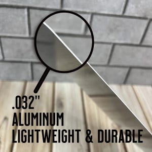 May include: A close-up of a silver metal sheet with the text ".032" inch aluminum lightweight & durable" printed on it. The sheet is being held up by a magnifying glass.