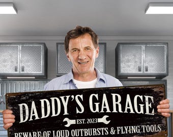 May include: A distressed black metal sign that reads "DADDY'S GARAGE" in white lettering, with the text "EST. 2023" and the warning "BEWARE OF LOUD OUTBURSTS & FLYING TOOLS". The sign is held by a smiling man.