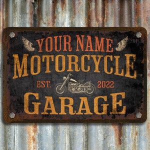 Pu&ograve; includere: Segno in metallo rustico con una silhouette di una motocicletta, il testo "YOUR NAME MOTORCYCLE GARAGE EST. 2022" e ali decorative.