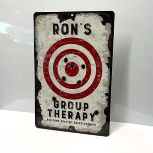 May include: A vintage-style metal sign with a red and white target with bullet holes. The sign reads "Ron's Group Therapy Building Healthy Relationships".