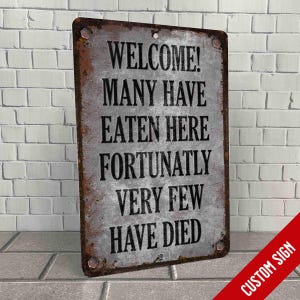 May include: A rusty metal sign with the text "WELCOME! MANY HAVE EATEN HERE FORTUNATELY VERY FEW HAVE DIED". The sign has a distressed, vintage look.