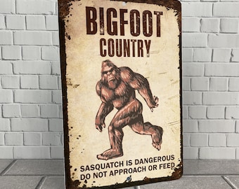 May include: A vintage-style metal sign with a distressed look. It features a cartoon illustration of Bigfoot walking, with the text "Bigfoot Country" and "Sasquatch is dangerous. Do not approach or feed."