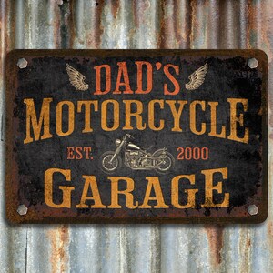 Pu&ograve; includere: Un cartello in metallo nero con una texture rugginosa. Il cartello recita "Garage di moto di pap&agrave; Est. 2000" con un'illustrazione di moto d'epoca al centro.