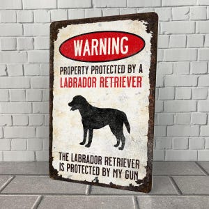 May include: A warning sign with a distressed metal look. The sign reads "WARNING PROPERTY PROTECTED BY A LABRADOR RETRIEVER THE LABRADOR RETRIEVER IS PROTECTED BY MY GUN". A black silhouette of a Labrador Retriever is in the center of the sign.