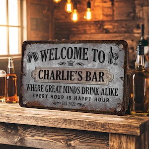 Peut inclure: Une enseigne métallique rouillée avec le texte "WELCOME TO CHARLIE'S BAR" et la phrase "WHERE GREAT MINDS DRINK ALIKE". L'enseigne comprend des illustrations de chopes de bière, une bouteille de vin et un verre. Sur une surface en bois.