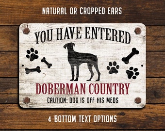May include: White sign with black text that reads "You Have Entered Doberman Country. Caution: Dog is off his meds."  The sign also features a black silhouette of a Doberman Pinscher dog, paw prints, and bone icons.
