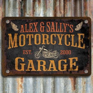 Pu&ograve; includere: Un cartello in metallo arrugginito con il testo "Alex & Sally's Motorcycle Garage Est. 2000" in lettere arancioni e nere. Il cartello presenta un'illustrazione in bianco e nero di una motocicletta con ali su entrambi i lati.
