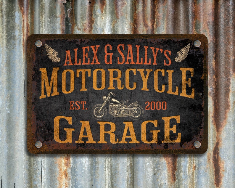 Pu&ograve; includere: Un cartello in metallo arrugginito con il testo "Alex & Sally's Motorcycle Garage Est. 2000" in lettere arancioni e nere. Il cartello presenta un'illustrazione in bianco e nero di una motocicletta con ali su entrambi i lati.