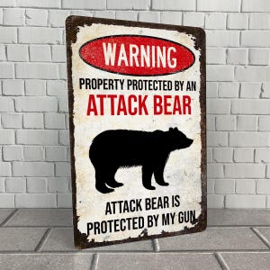 May include: A white metal sign with a red warning sign that says "WARNING" and "PROPERTY PROTECTED BY AN ATTACK BEAR". A black silhouette of a bear is shown below the text. The sign also says "ATTACK BEAR IS PROTECTED BY MY GUN".