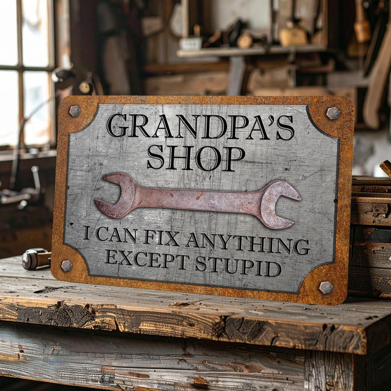 Puede incluir: Un letrero met&aacute;lico rectangular con un acabado envejecido. El letrero dice "GRANDPA'S SHOP" encima de un gr&aacute;fico de llave oxidada, con el texto "I CAN FIX ANYTHING EXCEPT STUPID" debajo. El letrero est&aacute; montado sobre una superficie de madera.