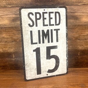 May include: A white metal sign with black text that reads "SPEED LIMIT 15". The sign has a distressed, vintage look.