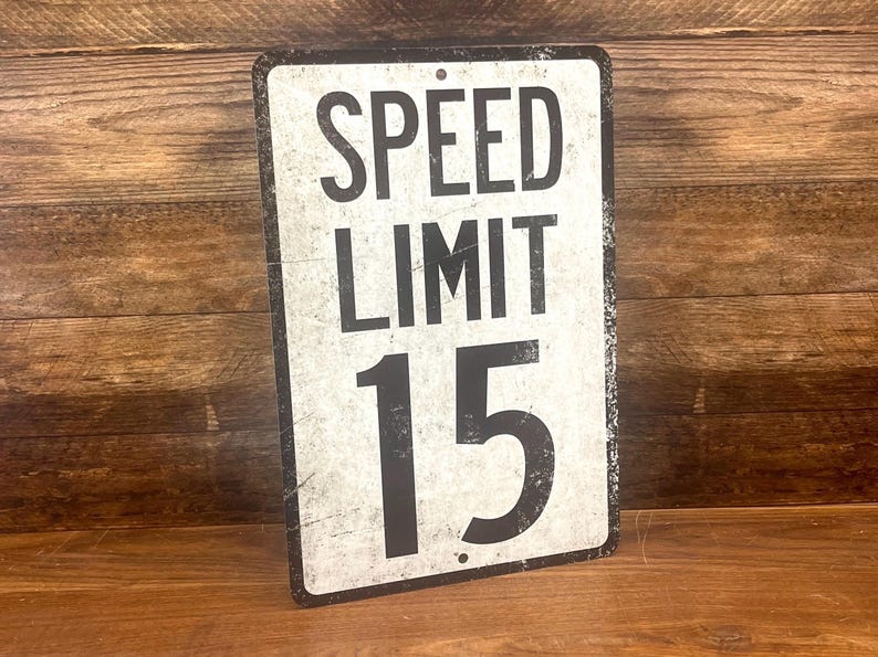 May include: A white metal sign with black text that reads "SPEED LIMIT 15". The sign has a distressed, vintage look.