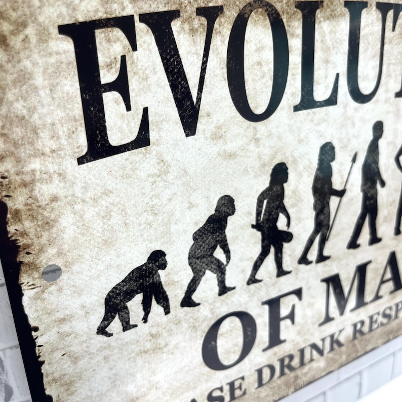 Op de afbeelding: Een vintage metalen bord met een zwarte achtergrond en witte tekst die "Evolutie van de mens" luidt, met een reeks silhouetten die de evolutie van de mens van aap tot moderne mens voorstellen. Het bord bevat ook de tekst "Drink verantwoord".