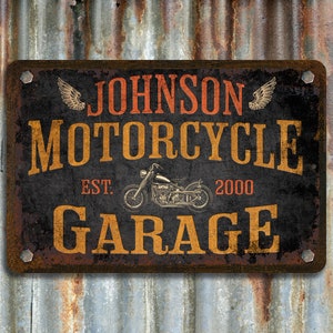 Pu&ograve; includere: Un cartello in metallo arrugginito con il testo "Johnson Motorcycle Garage Est. 2000" in lettere arancioni e gialle su sfondo nero. Il cartello ha una silhouette di una motocicletta e due ali su entrambi i lati del testo.