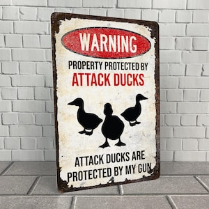May include: A white metal sign with a red warning sign that says "WARNING" and "PROPERTY PROTECTED BY ATTACK DUCKS". The sign also says "ATTACK DUCKS ARE PROTECTED BY MY GUN". The sign has a black silhouette of three ducks.