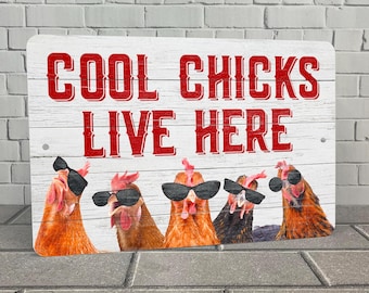 Op de afbeelding: Een wit houten bord met rode tekst die "Cool Chicks Live Here" luidt. Vijf kippen met zonnebrillen zijn te zien voor het bord.