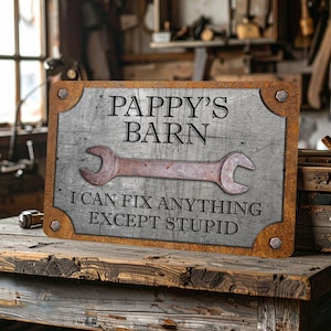 Puede incluir: Un letrero met&aacute;lico rectangular con el texto "PAPPY'S BARN" y "I CAN FIX ANYTHING EXCEPT STUPID". El letrero presenta una llave inglesa oxidada y una est&eacute;tica vintage y desgastada, ideal para un taller o garaje.