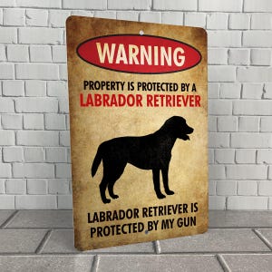 May include: A warning sign with a distressed, vintage look. The sign is brown with red and white text. It reads "WARNING PROPERTY IS PROTECTED BY A LABRADOR RETRIEVER LABRADOR RETRIEVER IS PROTECTED BY MY GUN". A black silhouette of a Labrador Retriever is in the center of the sign.