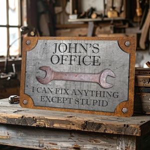 Puede incluir: Un letrero met&aacute;lico rectangular con un acabado envejecido, con el texto "JOHN'S OFFICE" sobre un gr&aacute;fico de llave inglesa. Debajo de la llave, el letrero dice "I CAN FIX ANYTHING EXCEPT STUPID". El letrero tiene una est&eacute;tica r&uacute;stica y vintage.