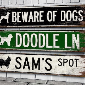 May include: Three wooden signs with distressed paint. The top sign is black with white text that reads "Beware of Dogs". The middle sign is green with white text that reads "Doodle Ln". The bottom sign is white with black text that reads "Sam's Spot".