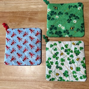 May include: Three quilted pot holders with loop handles. One is light blue with red lobsters, another is green with shamrocks and bees, and the third is white with shamrocks and bees. The pot holders are square and have a quilted pattern.