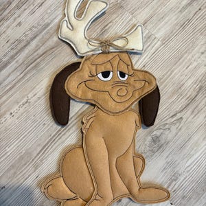 Peut inclure: Un personnage en feutre ressemblant à un chien, avec des oreilles marron et une expression triste, portant un serre-tête en forme de bois de cerf de couleur crème, noué avec de la ficelle. Le chien est assis, avec un corps brun clair et des contours brun foncé.