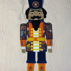 Könnte beinhalten: Dekorative Nussknacker-Puppe mit dem Logo der Houston Astros. Die Puppe hat einen marineblauen Hut, schwarze Haare und einen Bart sowie eine orangefarbene und marineblaue Uniform. Die Uniform hat goldfarbene Akzente und das Astros-Logo. Die Puppe ist etwa 61 cm groß.
