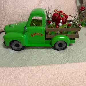 Könnte beinhalten: Ein hellgrüner Spielzeug-LKW aus Metall mit dem Wort GRINCH in roter Schrift auf der Tür. Die Ladefläche hat einen braunen Holzrahmen und ist mit einem roten Geschenk, grünem Lametta sowie kleinen roten und grünen Dekokugeln gefüllt.