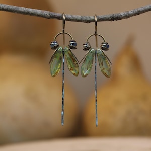 Può includere: Un paio di orecchini pendenti con ganci e cornici arcuate in metallo color bronzo. Ogni orecchino presenta due perline di vetro ovali, traslucide e verde pallido, e una lunga barra di metallo argentato sottile e testurizzata.