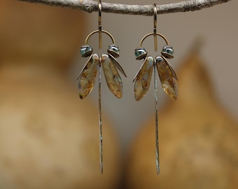 May include: A pair of dangle earrings featuring a brass arch wire, small metallic beads, and two elongated, mottled brown and gold-toned leaf-shaped metal charms hanging above a single, thin, textured silver-toned vertical metal bar.