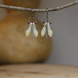 Puede incluir: Un par de pendientes colgantes con tres piezas en forma de pétalo de color blanco roto con vetas amarillas sutiles, suspendidas de una barra vertical de metal oscuro. Los pendientes incluyen pequeñas cuentas marrones y un arco de metal.