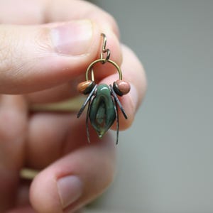 May include: A handcrafted earring featuring a green and brown teardrop-shaped bead, accented with small brown and pink beads. The earring has a brass-colored hoop and delicate metal details, held by a hand.