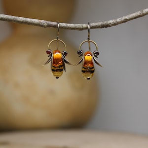 Op de afbeelding: Een paar handgemaakte oorbellen met een bijenontwerp. De oorbellen hebben een goudkleurig metalen frame met amberkleurige glazen lichamen en zwarte strepen. Kleine rode accenten zijn zichtbaar aan de bovenkant van elke oorbel. De oorbellen hangen aan een tak.
