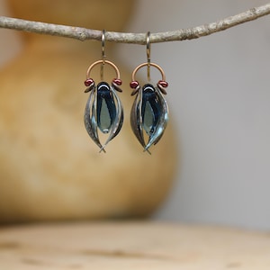 May include: Artisan earrings with a distinctive design. Each earring has a dark blue, teardrop-shaped glass bead within a silver-toned metal frame. They hang from copper-coloured hooks, adorned with small red beads, creating an elegant accessory.