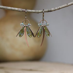 Op de afbeelding: Oorbellen in de vorm van een libel. Elke oorbel heeft een koperkleurig lichaam met groen-gouden vleugels en kleine blauwe kralen. De oorbellen hangen aan een kleine tak tegen een vage achtergrond.