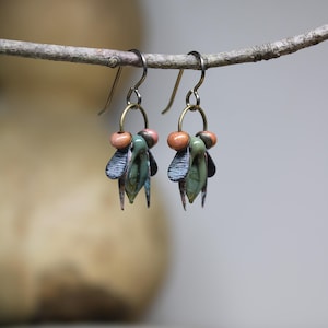 May include: Handmade earrings with a nature-inspired design. Each earring features a cluster of green, blue, and copper-colored elements, accented with small peach-colored beads. The earrings hang from simple, silver-toned hooks, creating a unique and artistic accessory.