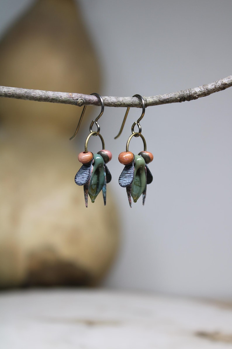May include: Handmade earrings with a nature-inspired design. Each earring features a cluster of green, blue, and copper-colored elements, accented with small peach-colored beads. The earrings hang from simple, silver-toned hooks, creating a unique and artistic accessory.
